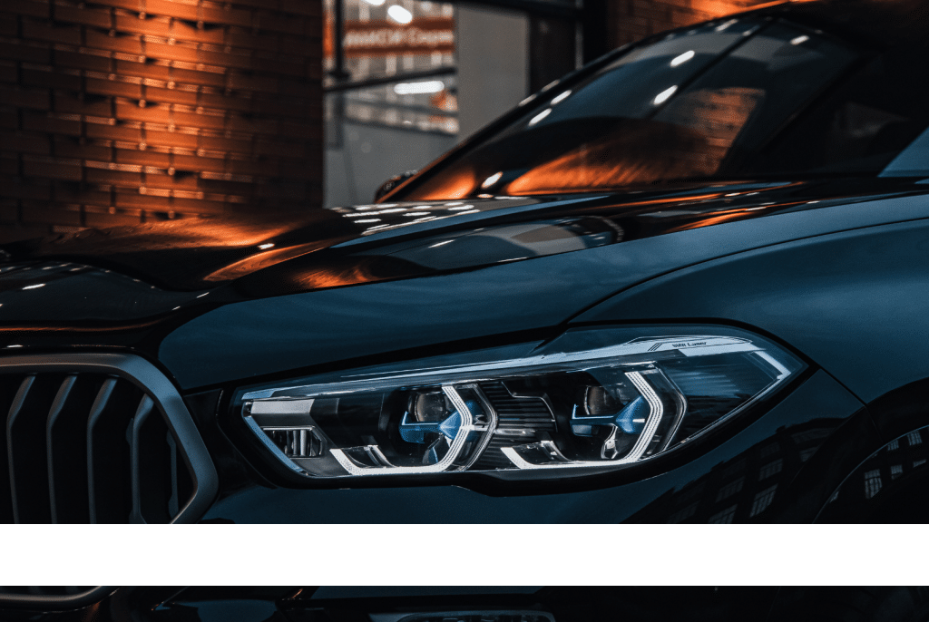 A close-up car photography image highlighting a luxury car's headlight and glossy reflections in an urban setting